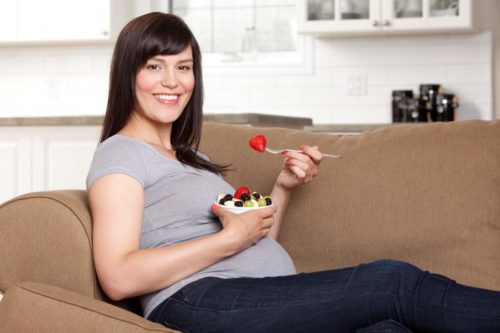אישה הריונית יושבת על ספה ואוכלת מקערה עם תותים וענבים טריים